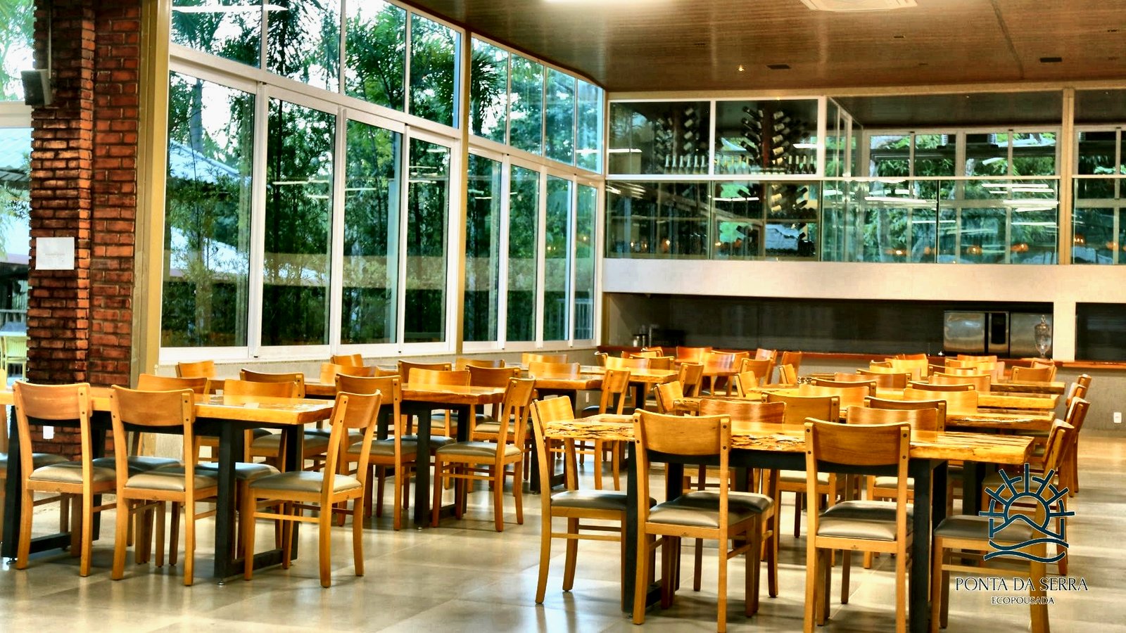 Salão do restaurante com mesas de madeira e grandes janelas para o verde